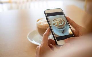 de hand av de kvinna använder sig av en smartphone till ta en bild av en kaffe i en Kafé till posta på social media foto