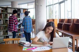 ung asiatisk studenter i tillfällig ha på sig do läxa och använda sig av en teknologi bärbar dator i de universitet bibliotek. de studenter är sökande för kunskap och är framställning för de examen. foto