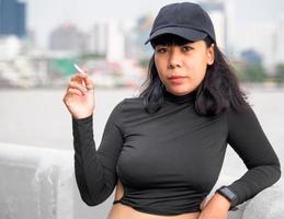 porträtt kvinna flicka tonåring ung Asien ett person bär en hatt och lång svart hår bär en svart skjorta hand håll rökning cigarett vit Färg stående utomhus- förbi de vägg utomhus- foto