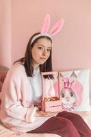 en skön ung flicka med kanin öron på henne huvud sitter på Hem på en säng med en korg av dekorerad påsk ägg. förberedelse för påsk. foto