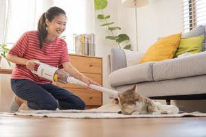 Lycklig asiatisk ung hushållerska kvinna använder sig av Vakuum rengöring, rengöringsmedel till ta bort damm, hår eller päls på golv i levande rum medan söt katt liggande på matta. rutin- hushållsarbete, Syssla i hushåll av hembiträde. foto