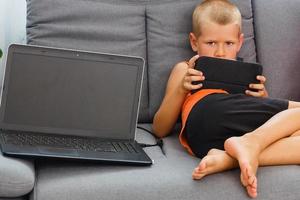 liten pojke med digital läsplatta Sammanträde på soffa, på Hem interiör bakgrund foto