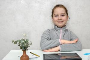 liten söt flicka studerande med en leende sitter på en skrivbord med henne händer knäppt redo för klasser, tillbaka till skola foto