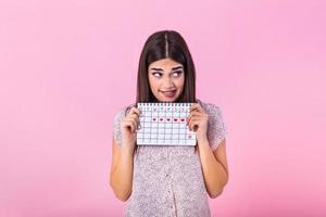 porträtt av generad söt flicka innehav menstruations- kalender med dragen hjärtan för period och ser på kamera isolerat över rosa bakgrund foto