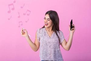 kvinna bär hörlurar lyssnande till musik från smartphone studio skott isolerat på rosa bakgrund med musik anteckningar. upphetsad ung kvinna har roligt i hörlurar med telefon på rosa studio bakgrund. foto