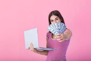 bild av glad ung kvinna stående isolerat över rosa bakgrund använder sig av bärbar dator dator och innehav pengar sedlar . porträtt av en leende flicka innehav bärbar dator dator foto