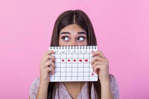 porträtt av en rolig ung flicka i dölja Bakom en menstruations- perioder kalender och ser bort på kopia Plats isolerat över rosa bakgrund. kvinna period kalender foto