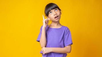 ung asiatisk tjej i lila skjorta som pekar uppåt och tänker i studio med gul bakgrund