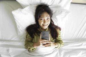 en leende ung asiatisk kvinna är skrattande medan innehav henne telefon och liggande på de säng foto