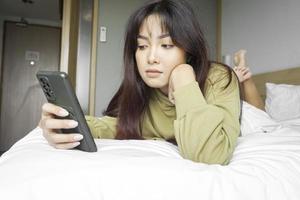 en ung asiatisk kvinna är känsla allvarlig och fokuserade på innehav henne smartphone i hand medan liggande på de säng. foto