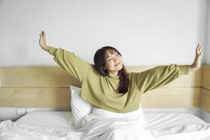 porträtt av skönhet Lycklig asiatisk kvinna vakna, vakna upp på de säng. ung skön asiatisk flicka stretching henne vapen i färsk tidigt morgon, koppla av fredlig morgon- rutin- gäspa begrepp baner foto