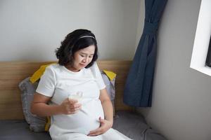Lycklig ung asiatisk gravid kvinna dricka mjölk på säng på Hem medan innehav henne mage och tänkande foto