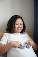 ung Lycklig asiatisk gravid innehav små skor för de ofödd bebis i henne hand och se in i de kamera foto