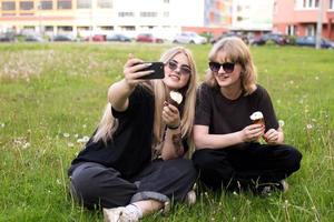 två rolig flickor är äter is grädde och tar selfies på de gata. tonåringar i social nät foto