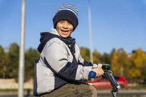 en glad tandlös preschooler pojke ler och rider en cykel i ett höst hatt och jacka foto