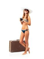 full längd porträtt av en skön ung kvinna Framställ i en bikini med en kamera i händer och resväska på vit foto