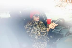 skön kvinna i en päls täcka och röd hatt med kaffe till gå körning henne bil foto