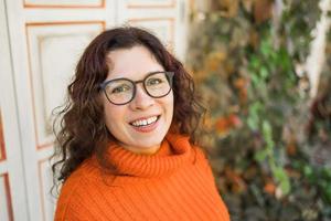 Lycklig porträtt av attraktiv ung kvinna i eleganta glasögon i stickat modern orange Tröja på bakgrund av löv kopia Plats och tömma Plats för text. flicka promenader i stad foto