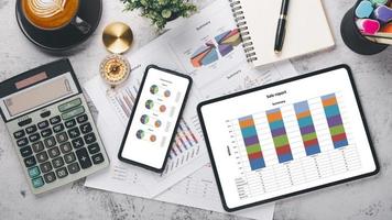 läsplatta och smart telefon med diagram och rapporter, kalkylator, penna, anteckningsbok och kaffe på kontor skrivbord arbetsplats. topp se platt lägga foto