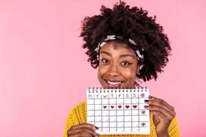 porträtt av leende ung afrikansk amerikan kvinna innehav period kalender isolerat över rosa bakgrund. skön ung kvinna stående isolerat över rosa bakgrund, som visar menstruations- kalender foto