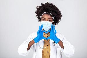 kvinna afrikansk professionell läkare sjuksköterska, läkare ha på sig ansikte mask, handskar, enhetlig som visar hjärta händer form. medicinsk kärlek, vård och säkerhet symbol, korona virus hälsa skydd tecken begrepp. närbild foto
