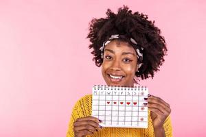 porträtt av leende ung afrikansk amerikan kvinna innehav period kalender isolerat över rosa bakgrund. skön ung kvinna stående isolerat över rosa bakgrund, som visar menstruations- kalender foto