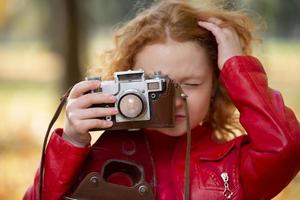 liten rödhårig flicka med en retro kamera på ett höst bakgrund. foto