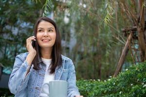 ung asiatisk kvinna frilansare innehav smartphone för talande med vän. livsstil kvinnor arbetssätt på Hem begrepp. Sammanträde i snår av tropisk. flicka på Semester Sammanträde i trädgård leende avkopplande. foto