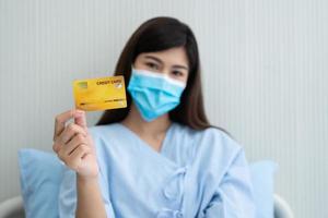glad asiatisk kvinna som bär en medicinsk mask och håller mock up kreditkort försäkringskort och ok tecken i en sjukhussäng. försäkring av bank, betalning medicinsk behandling koncept foto