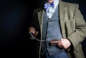 porträtt av brittiskt herre i tweed kostym och läder handskar innehav guld ficka Kolla på på svart bakgrund. retro stil och årgång mode. foto