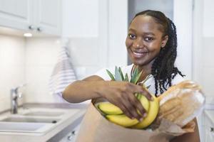 friska positiv Lycklig kvinna innehav en papper handla väska full av frukt och grönsaker. ung kvinna innehav matvaror handla väska med grönsaker stående i de kök foto