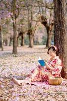 ung lady i traditionell kimono klänning läsning bok på körsbär blomma träd. känsla leende foto