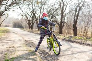 7 år gammal Lycklig liten flicka barn rida cykel i de parkera på Hem och bär skydd mask för skydda PM 2,5 och coronavirus covid-19 pandemi virus symptom.sport övning för hälsa. foto