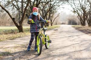 7 år gammal Lycklig liten flicka barn rida cykel i de parkera på Hem och bär skydd mask för skydda PM 2,5 och coronavirus covid-19 pandemi virus symptom.sport övning för hälsa. foto