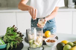 ung smal kvinna i vit t-shirt och blå jeans matlagning frukt smoothie med frön friska mat i kök på Hem foto