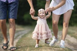 Lycklig ett år gammal flicka är varelse hölls förbi pappa och mamma. liten barn gående med föräldrar. en söt bebis lär till promenad med de hjälp av henne föräldrar. foto
