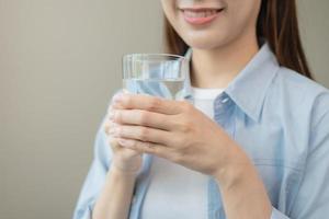 Lycklig skön, asiatisk ung kvinna, flicka dricka, smutta färsk glas av vatten för hydratisering av kropp, innehav transparent glas i henne hand, törstig på Hem. hälsa vård, friska livsstil begrepp. foto