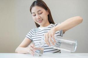 Lycklig skön, törstig asiatisk ung kvinna, flicka dricka, hand i innehav, häller vatten från kanna in i transparent glas från tillbringare för hydratisering av kropp. hälsa vård, friska livsstil begrepp. foto