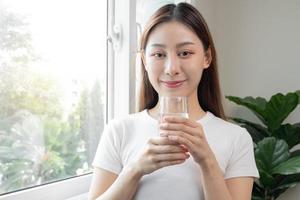 Lycklig skön, asiatisk ung kvinna, flicka dricka, smutta färsk glas av vatten för hydratisering av kropp, innehav transparent glas i henne hand, törstig på Hem. hälsa vård, friska livsstil begrepp. foto