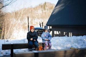 bror med syster Sammanträde förbi läger bål på vinter- mot mycket liten hus. barn i landsbygden. foto