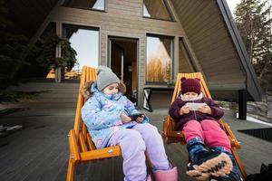 två liten flickor sitta på stolar på terrass av rutnät mycket liten hus i de bergen och tittar på tecknade serier på mobil telefoner. foto