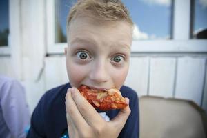 Foto vid vinkel lins av en liten pojke vem äter pizza med hans ögon bred öppna.