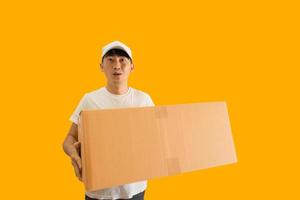 ung asiatisk leverans man bär keps och vit tom t-shirt innehav paket posta låda isolerat på gul bakgrund. uttrycka leverans service begrepp. foto