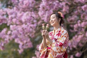 japansk kvinna i traditionell kimono klänning innehav ljuv hanami dango efterrätt medan gående i de parkera på körsbär blomma träd under vår sakura festival med kopia Plats foto