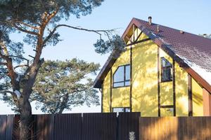 en gul mysigt hus i de snö i vinter- i de by är omgiven förbi tall träd. snötäckt tak, uppvärmning och ventilation rör, trapezoid fönster foto