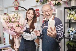 e-handel företag, senior asiatisk manlig blomsterhandlare arbetstagare och dotter visa blommig arrangemang, uppkopplad leva strömning, selfie förbi smartphone Ansökan i ljus blomma affär, skön blommar Lagra. foto