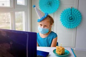 en Lycklig liten flicka, bär en medicinsk mask i främre av henne bärbar dator firar henne födelsedag via de internet i karantän tid, självisolering, uppkopplad födelsedag. foto