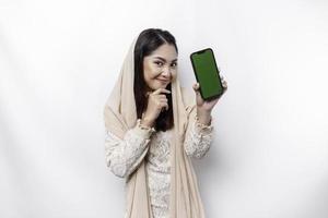 en omtänksam ung asiatisk muslim kvinna bär en hijab och innehav henne haka medan som visar de telefon skärm isolerat förbi en vit bakgrund foto