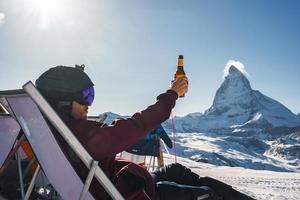 ung snowboardåkare glädjande med en öl efter skidåkning dag i en bar eller en Kafé på de zermatt åka skidor tillflykt i swiss alps med en fantastisk se av de matterhorn. vinter- semester begrepp. foto