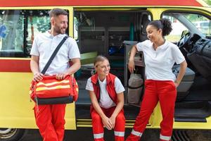 paramedics och läkare stående på de sida ambulans. läkare är bärande en medicinsk trauma väska. grupp av tre paramedics stående i främre av ambulans med leende. foto